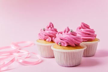 A beautiful cupcakes with bright pink cream and a little silver decoration on a pink background. Pink ribbon for decor in the foreground lies in waves. Festive background with free space.