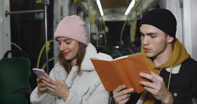 Millennial guy using reading book while sitting near his lovely girlfriend. Young pretty girlusing smartphone while going on public transport with her boyfriend. Concept of real life.