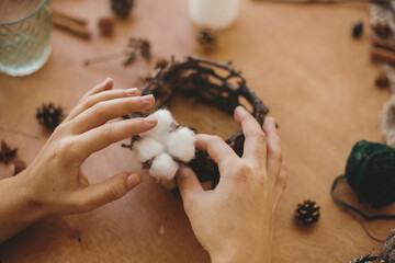 Florist making rustic christmas candle holder. Hands holding cotton and making rustic festive decoration on wooden table with branches and pine cones. Holiday workshop