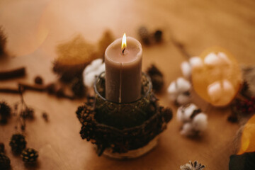 Christmas rustic candle in festive lights bokeh on wooden table with pine cones, cotton,anise and branches. Cozy moody winter evening. Rustic christmas candle decor on table.