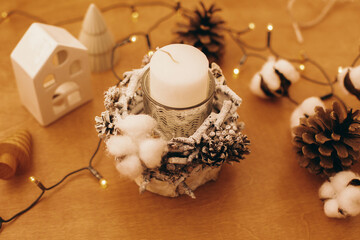 Christmas candle in rustic holder made of branches, cotton, anise and pine cones on wooden table with festive lights. Holiday workshop. Making rustic christmas candle decor.