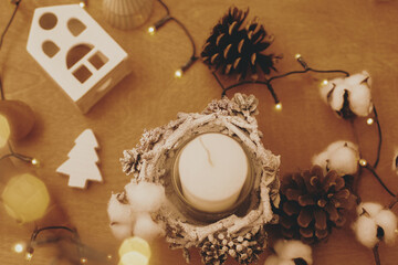 Christmas rustic candle, pine cones, wooden tree, toy house and  festive lights on wood. Flat lay. Rustic christmas candle decor on table. Winter holiday