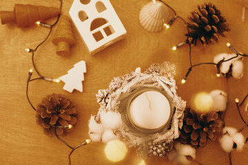Christmas rustic candle, pine cones, wooden tree, toy house and  festive lights on wood. Rustic christmas candle decor on table. Winter holiday
