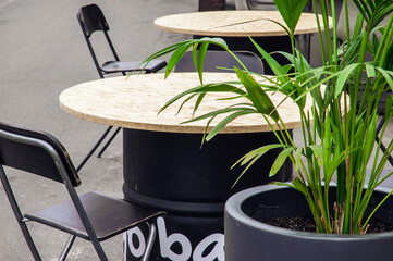 Summer terrace bar with modern metallic barrel tables with round counter and wooden bar chairs. Reastaurant tables near green pam shrub waiting for customers on city street