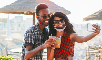 African american couple taking selfie in beach party - Young friends with face mask drinking...