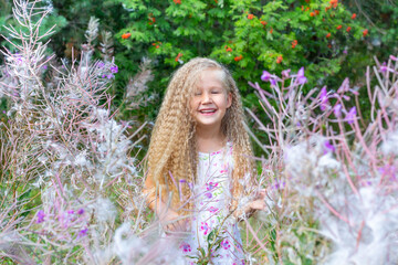 A little blonde girl of 5-6 years old in a white sundress stands surrounded by blooming Sally, fireweed. Long blonde curly hair, hairstyle. Summer, nature, lifestyle. Emotions, smile, happy childhood.