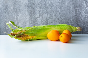 An ear of corn and three apricots lie on a white table against the background of a gray concrete wall. Ecology concept of vegetarianism. Healthy food, ingredients, fruits and vegetables.
