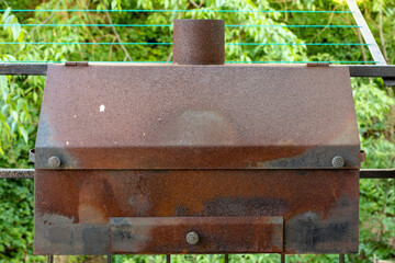 Fototapeta na wymiar Old rusty large size barbecue grill with ash tray and chimney hanging on iron bars with trees in the background.