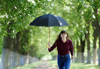 A girl with an open black umbrella runs along the road in the crowns of green trees.
