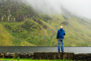 Fototapeta na wymiar Bald male tourist enjoy view in Connemara region, Ireland. Man standing on a stone fence by a lake, Magnificent mountain covered with clouds in the background. Wild Atlantic way route.