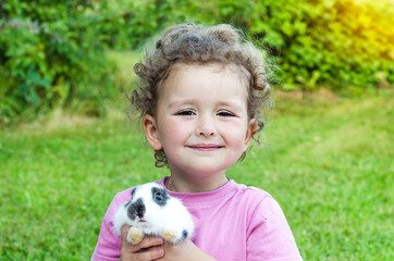 little beautiful girl smiling, hugging a baby rabbit, hamster on the green grass in summer. happy laughing child and pet outdoors. bunny is a symbol of Easter. soft focus
