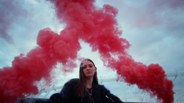 Girl posing at camera in colorful smoke. Attractive woman holding smoke bombs