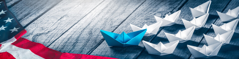 Blue Paper Boat Leading A Fleet Of Small White Boats With American Flag On Wooden Table -...
