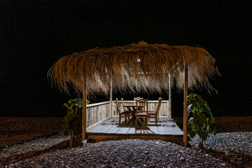 Cozy gazebo by the sea. Thatched roof, bamboo railing, wooden furniture. Windy evening in a cafe,...