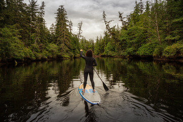 Girl Paddle Boarding in a River near the Pacific Ocean with green trees during a cloudy day. Taken in Raft Cove, Northern Vancouver Island, British Columbia, Canada.