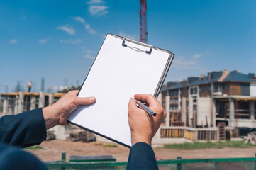 The chief in a jacket and a construction helmet with documents in his hands, against the background of a tower crane and the sky with clouds. House construction inspection.