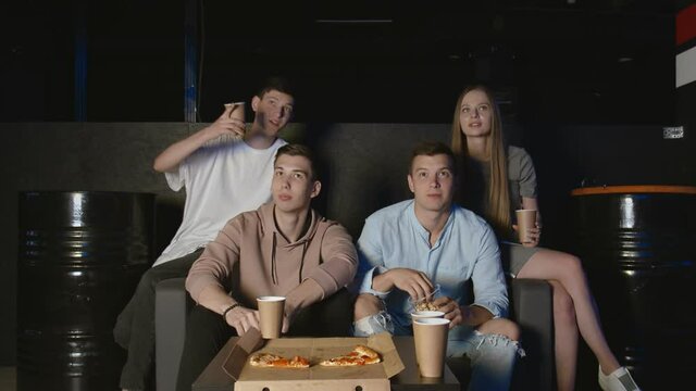 Group of young friends watching television together on couch, communicating, eating popcorn and pizza