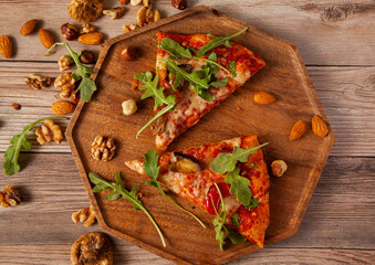 Flat lay close up image of  vegetarian cheese and tomato pizza decorated with fresh arugula leaves and grilled egg plants slices. It is served on wooden tray with hazelnuts, almonds and walnuts