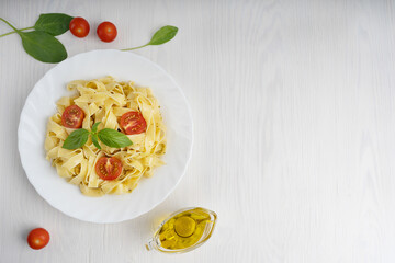 Top view of organic italian pasta decorated with sliced tomatoes and basil leaves surrounded by olive oil gravy boat and spinach on white wooden background at kitchen. Image with copy space