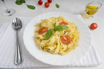 Traditional orgabic italian fettucine pasta decorated with sliced tomatoes and basil leaves in a plate served together with spoon and fork on towel on white wooden background at cafe