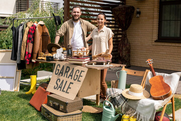 Portrait of smiling young couple standing at table with Garage sale sign and selling stuff in backyard