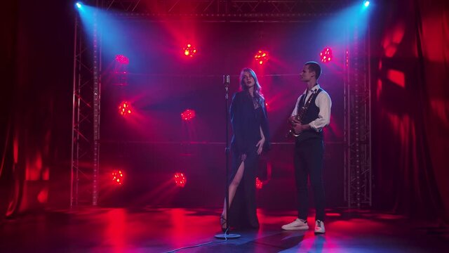 A young girl singer looks passionately at a guy saxophonist and touches it with his hand. A brunette in a long black dress and men jacket. Dark studio with smoke and neon lighting. Dynamic red neon