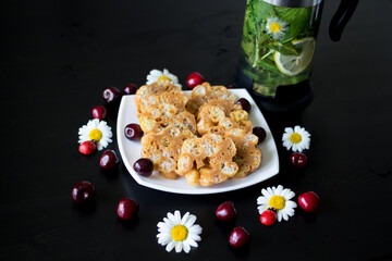 Dessert of toffee and corn sticks on a white square plate, decorated with daisies with ladybirds and cherries. Next to the kettle, which is brewed currant leaf, lemon, mint and chamomile...