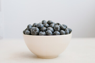 Minimalistic photo of freshly picked sweet blueberry in ivory bowl on dark blue background. Handful of sweet ripe berries close up 