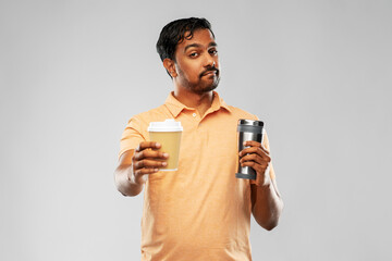 eco living and sustainability concept - young man in polo t-shirt comparing thermo cup or tumbler with disposable paper coffee cup over grey background