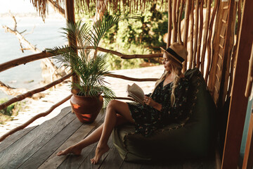 Young happy tourist woman in straw hat sit on the balcony of wooden eco  bungalow with sea view and read a book