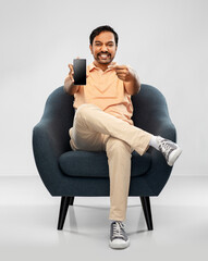 people, technology and furniture concept - happy smiling young indian man showing smartphone...