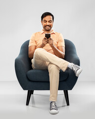 people, technology and furniture concept - happy smiling young indian man with smartphone sitting...