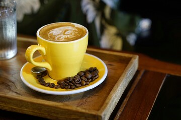 A yellow cup of cappucino coffee with beans on wooden tray and table in coffee shop, green vintage background.