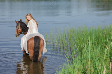 Young blonde girl in a white long dress swims in the lake with a brown horse on a sunny day
