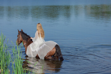 Young blonde girl in a white long dress swims in the lake with a brown horse on a sunny day