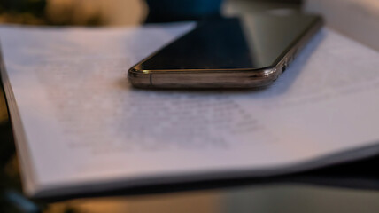 Open book and phone on a glass black table. Blurred boke background, close-up. Dark evening background with sunset highlights.