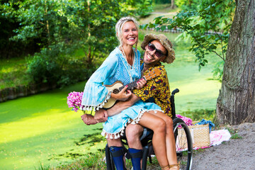 blond woman with guitar sitting on lap of man in wheelchair outdoors in park and hugging him
