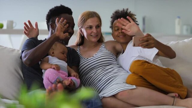 Young multiracial family with children taking selfie photo on couch in apartment interior spbi. Dad, mom and two kids having fun and making picture sitting on sofa in light room. Happy multiethnic