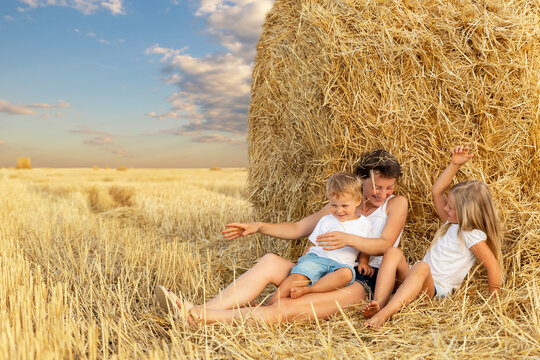 Young adult attractive beautiful mom with little son and daughter enjoy having fun fooling around sitting near golden hay bale on wheat harvested field near farm. Happy children on rural landscape