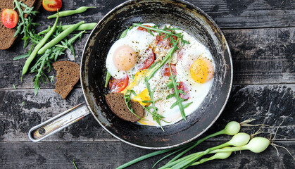 Rustic breakfast. Fried eggs with vegetables: tomatoes and asparagus beans in a pan with bread, arugula, onions on old wooden black background in nature. Summer, outdoor. Background image, copy space