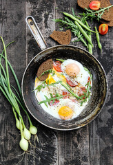 Rustic breakfast. Fried eggs with vegetables: tomatoes and asparagus beans in a pan with bread, herbs, onions on old wooden black background in nature. Summer, outdoor. Background image, copy space