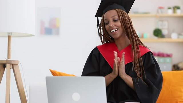 African American female student talks and claps hands in front of laptop screen in apartment spbi. Young attractive person having distant conversation with smile and using computer, sitting at table
