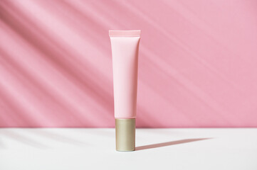 Pink tube of soft organic natural eye cream with golden cap on white pink background, palm leaf shadow. Summer facial product, sun lotion. Women's hygiene cosmetic accessory for skincare. Copy space.