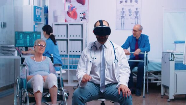 Doctor wearing VR headset, diagnosing senior woman problems while she sitts in a wheelchair in the back. Futuristic medicine, health care of the future in private clinic or hospital