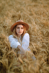 Naklejka na ściany i meble Young woman in white linen dress and hat enjoying a sunny day in a golden wheat field. Summer, beauty, fashion, glamour, lifestyle concept.