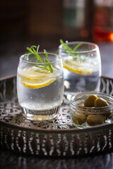 2 glasses of gin & tonic with bowl of olives on a drinks tray.  Drinks garnished with rosemary lemon and ice.