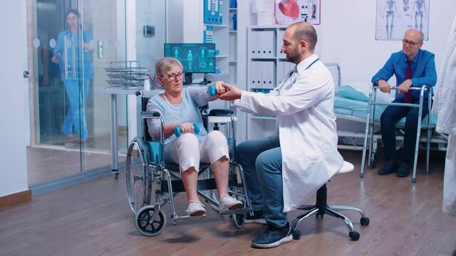 Doctor helping old disabled woman in wheelchair to regain muscle strength in a private recovery clinic. Invalid person using dumbbells to exercise. Rehabilitation hospital, working with paralyzed