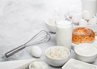 Fresh dairy products on white table background. Glass of milk, bowl of sour cream and cottage cheese and eggs in wooden box. Fresh baked baggel on round chopping board. Steel whisk. Space for text