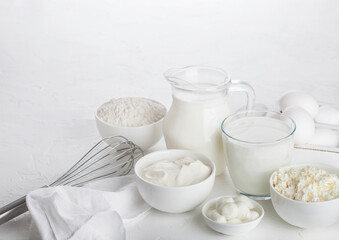 Fresh dairy products on white table background. Jar and glass of milk, bowl of sour cream, cottage cheese and baking flour and mozzarella. Eggs and cheese. Steel whisk