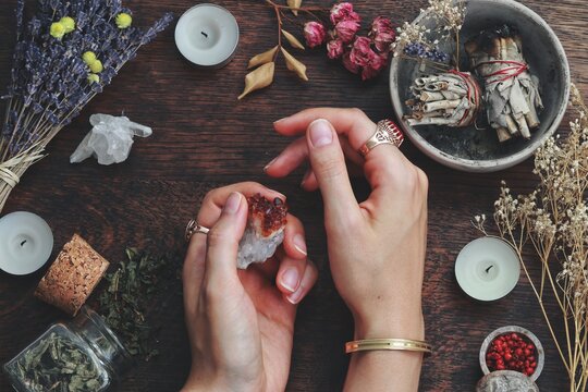 Witches hands on a table ready for spell work. Wiccan witch altar filled with sage smudge sticks, herbs, white candles. Female witch wearing vintage jewelry, holding citrine crystal rock in her hands